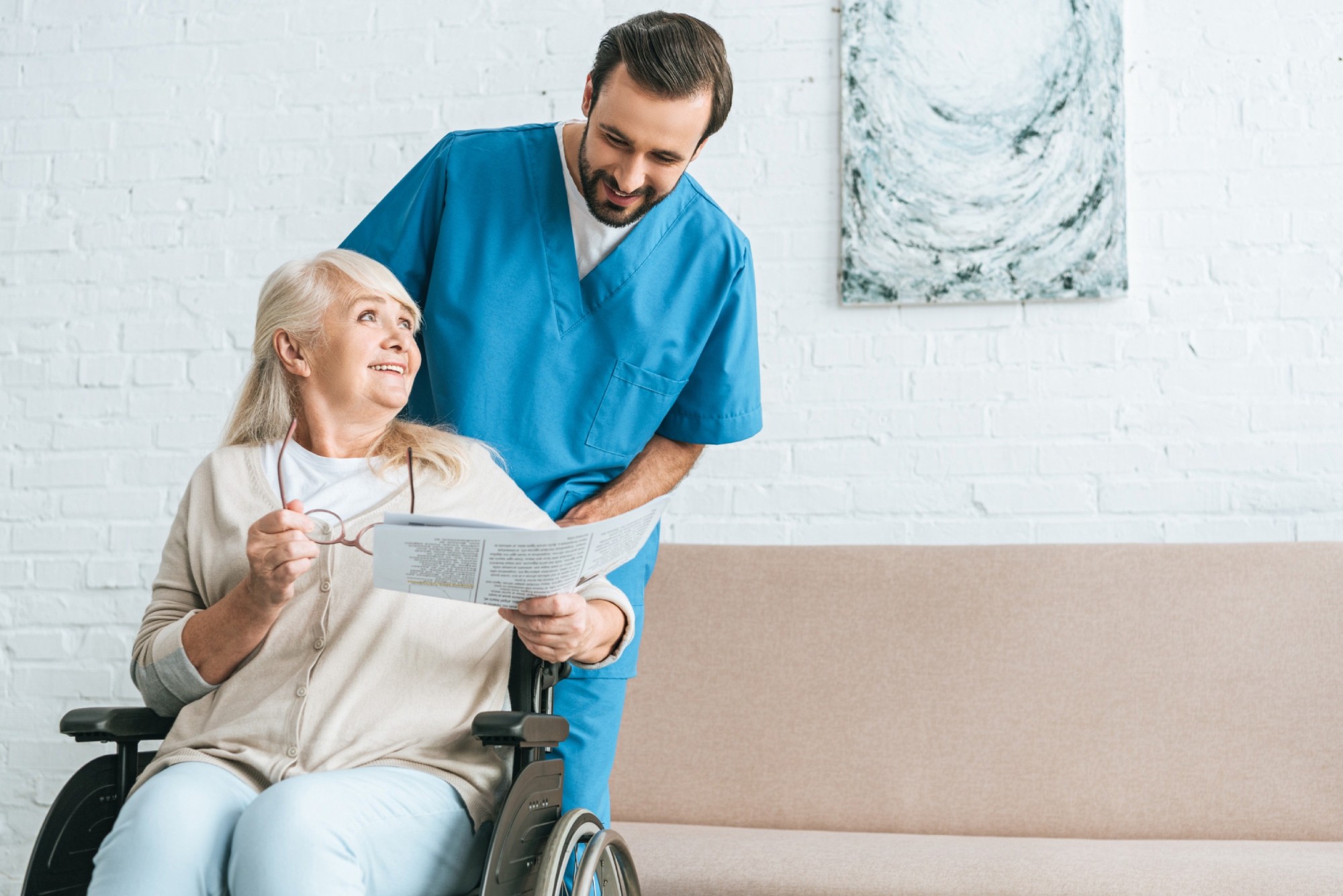 smiling young male nurse looking at happy senior woman reading newspaper in wheelchair