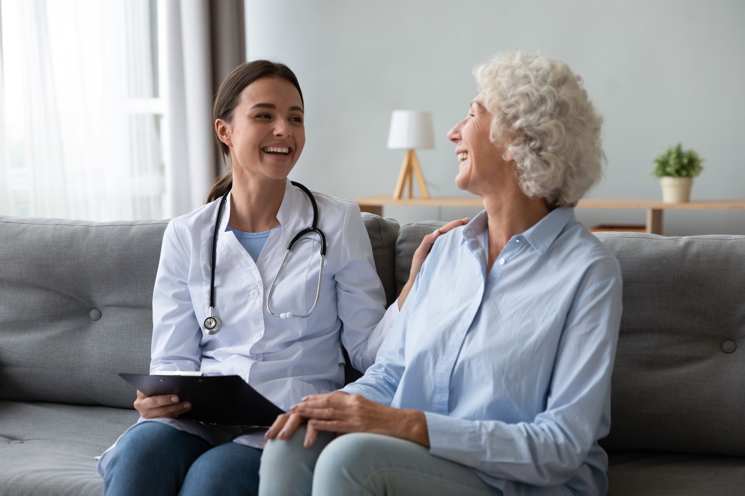 Friendly young nurse in white coat holding clipboard laughing with elderly female patient during homecare visit consultation, provide her support encourages aged woman enjoy warm talk seated on couch